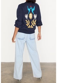 Marinblå tröja med färgglad broderi av fåglar, hjärtan och löv på ryggen; ihop med ljusblå vida jeans med scallopformade fickor.