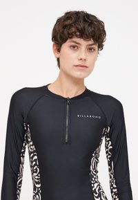 Black long-sleeve rash guard with a front zipper, featuring a white floral design on the sides and a "BILLABONG" logo on the chest.
