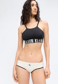 Γυναίκα με μαύρο bralette Calvin Klein και λευκά μαγιό κάτω μέρος Calvin Klein, στέκεται μπροστά σε απλό φωτεινό φόντο.