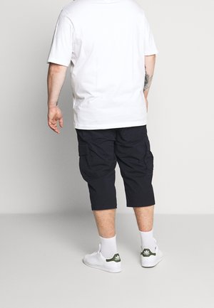 Person iført hvid t-shirt, sorte cargoshorts, hvide sokker og hvide Adidas-sneakers, stående med ryggen til på en enkel baggrund.