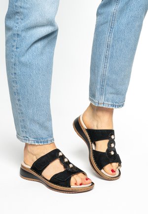 Füße in hellgewaschenen Jeans und schwarzen offenen Sandalen mit Perlendekor und gepolsterten Sohlen auf weißem Hintergrund.
