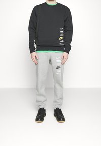 Person som bär en svart sweatshirt och grå sweatpants med flera Nike-logotyper, svarta sneakers och en liten axelväska.