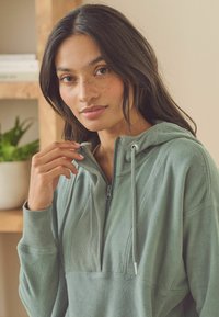 Fleece hættejakke i dæmpet grøn med en halvlynlås foran, snore og ribbede detaljer på ærmerne og skuldrene. Glat tekstur.