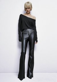 Top nero senza spalline con maniche lunghe, abbinato a pantaloni in ecopelle a vita alta e svasati con dettagli decorativi a occhiello e cerniere.