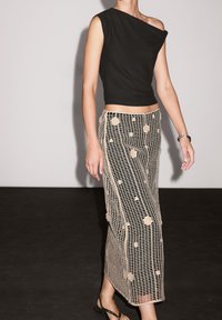 Top noir à épaule unique associé à une jupe en crochet beige avec des motifs circulaires. La tenue est complétée par des sandales plates noires.