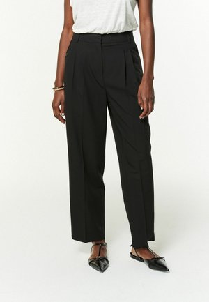 Zwarte wijde broek met een geplooid voorpand, hoge taille en knoopdetails, gecombineerd met zwarte puntige flats.