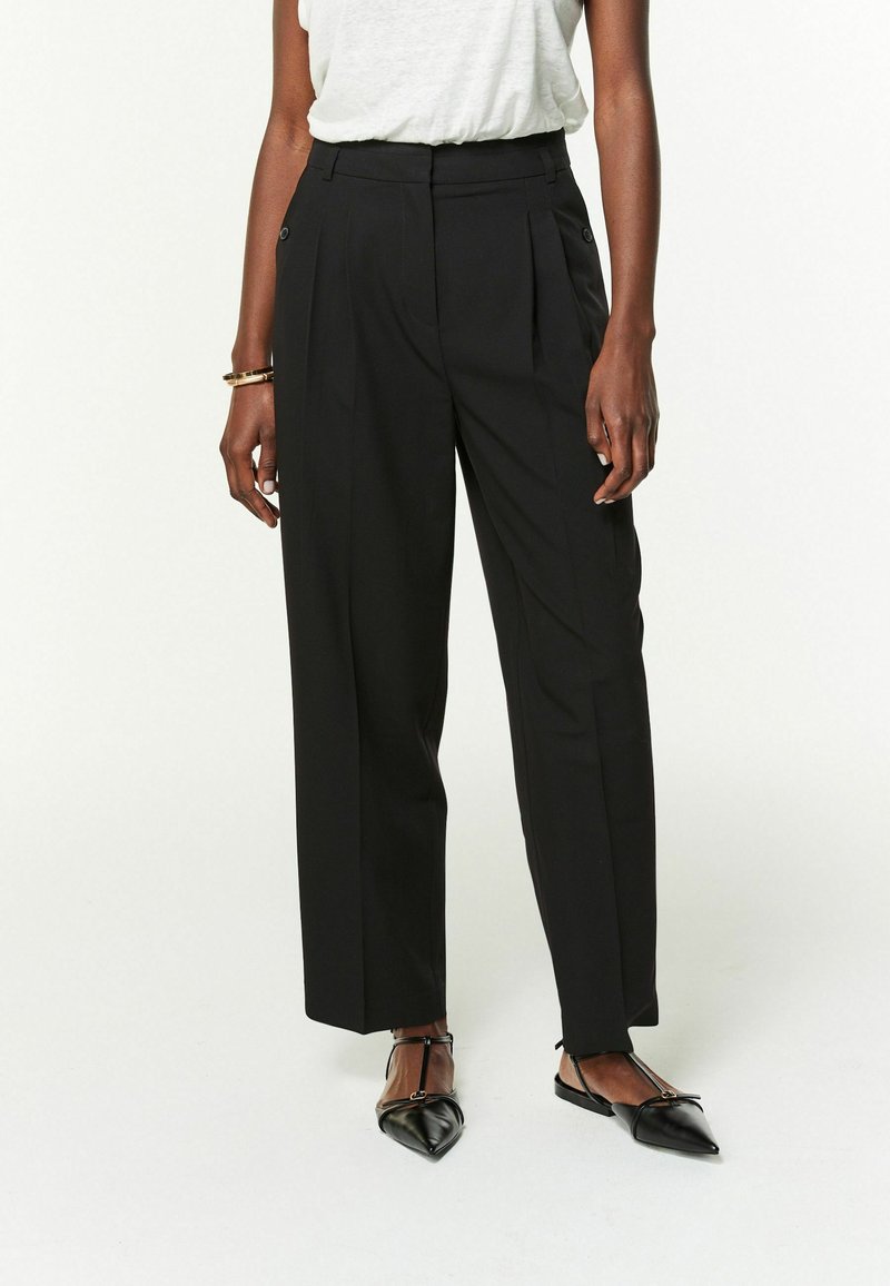 Pantalons larges noirs avec un devant plissé, taille haute et détails en boutons, associés à des chaussures plates noires à bout pointu.