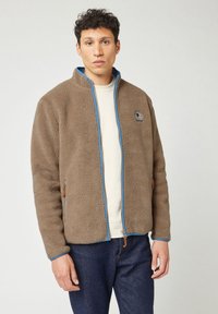 Giacca in pile marrone con accenti blu, zip completa, due tasche laterali e logo ricamato. Indossata sopra una camicia di colore chiaro.