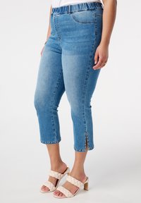 Jeans capri en denim bleu avec un design ajusté, une taille élastique, des fentes latérales avec des accents métalliques et une texture lisse. Associés à des sandales à talons beiges.