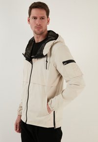 Lichtbeige, lichtgewicht jack met een trekkoordcapuchon, ritszak op de borst, elastische manchetten en een ritssluiting aan de voorkant. Gevoerde zwarte binnenkant.