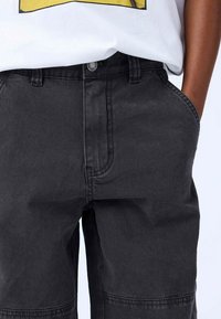 Shorts in denim nero con una vestibilità morbida, dotati di tasche laterali, chiusura con zip frontale e una leggera texture sbiadita. Vita regolabile visibile.