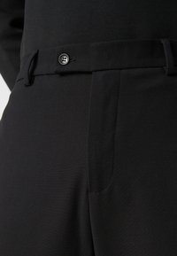 Pantalon noir en tissu texturé avec une poche latérale, des passants de ceinture et une fermeture à bouton à la taille. Design épuré et ajusté.