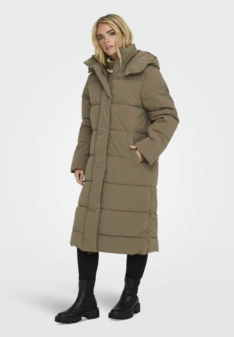 Manteau d'hiver long, rembourré, vert olive avec une capuche, présentant un design matelassé horizontal et des poches avant. Porté avec un jean skinny noir et des bottes.