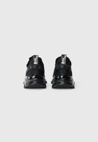 Une paire de baskets noires slip-on avec des semelles texturées et des tirants au talon portant le texte de la marque, vues de l'arrière sur un fond blanc.