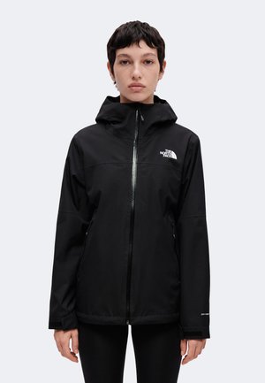 Osoba ubrana w czarną kurtkę North Face z kapturem i zamkiem błyskawicznym, stojąca przodem na jednolitym jasnym tle.