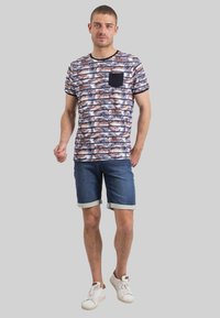 Chemise à manches courtes avec motif de feuilles de palmier en marine, orange et blanc ; présente une poche marine. Assortie à un short en denim et des baskets blanches.