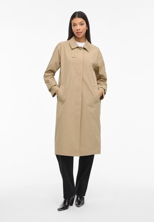 Manteau beige avec un seul bouton à l'avant, col pointu et poches latérales. Tissu lisse et design mi-long. Porté sur un pantalon noir.