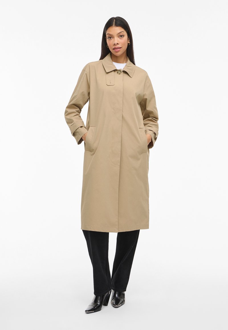 Impermeabile beige con un unico bottone frontale, colletto a punta e tasche laterali. Tessuto liscio e design di lunghezza intermedia. Indossato sopra pantaloni neri.