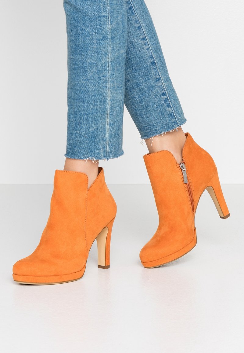 Femme portant des bottines en daim orange vif à talons hauts avec fermeture éclair latérale et jean bleu raccourci sur un sol blanc.