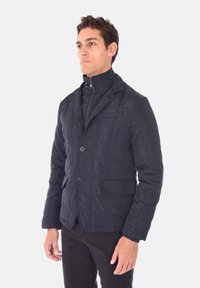 Giacca trapuntata blu navy con colletto alto, due tasche frontali e chiusura a un bottone, caratterizzata da una texture di tessuto liscia e lucida.