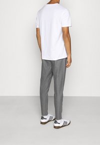 Mand iført hvid kortærmet t-shirt, grå bukser, hvide strømper og hvide sneakers med sorte striber, der står mod en ensfarvet lys baggrund.