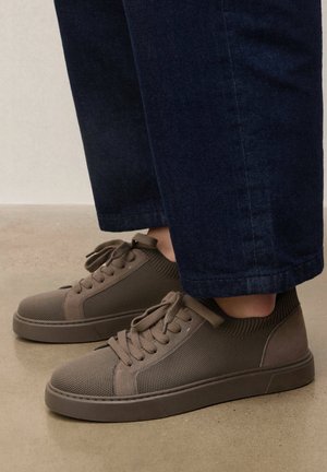 Személy barna kötött alacsony szárú cipőben, fűzővel, sötétkék rövid szárú nadrágban, sima bézs padlón állva.