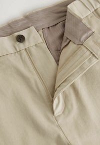 Beige Hosen aus glattem Stoff mit einem Reißverschluss und Knopfverschluss. Innen sind sie mit einem kontrastierenden, seidigen Futter versehen. Sichtbare Nähte.