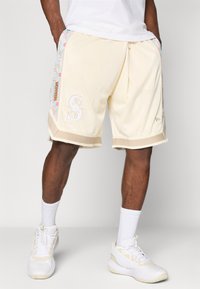 Calções de malha cremosos com painéis laterais florais, apresentando um acabamento em bege contrastante e detalhes em branco do logótipo na perna esquerda.