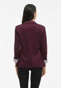 Femeie cu păr lung și închis la culoare, îmbrăcată într-un sacou elegant de culoare prună, cu manșete rayate și pantaloni negri, stând cu spatele îndreptat spre un fundal alb.