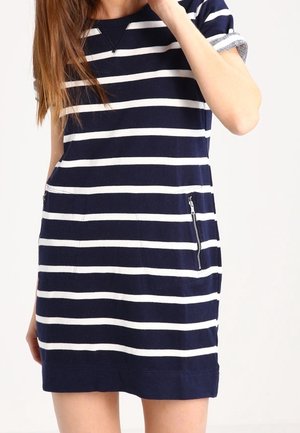 Kobieta w granatowej sukience z białymi poziomymi paskami, krótkimi rękawami i kieszeniami na suwak po bokach z przodu.