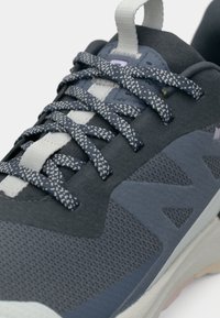 Αθλητικό παπούτσι σκούρου γκρι χρώματος με υλικά δίχτυ και σουέτ, υφή με ανάγλυφο φινίρισμα, αντίθετες λευκές λεπτομέρειες και κορδόνια με σχέδια.
