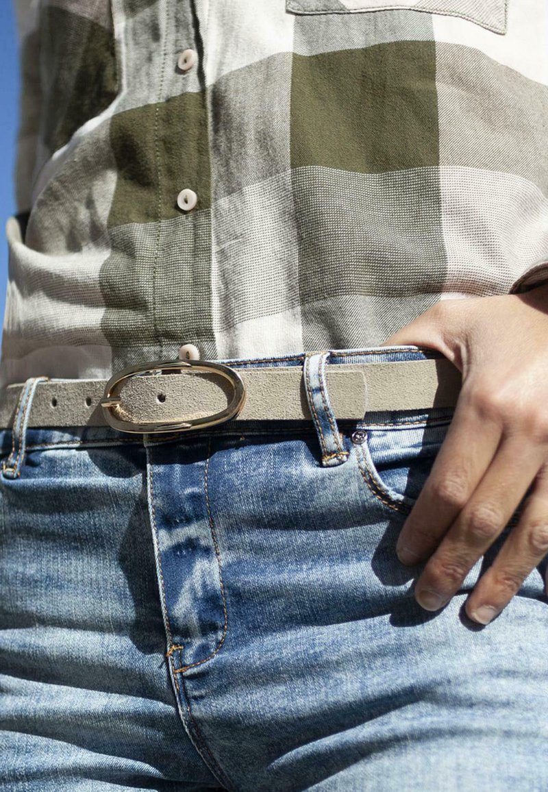 Cinturón de cuero gris claro con una hebilla ovalada en tono dorado, usado sobre unos pantalones de mezclilla azules. La camisa presenta un patrón de cuadros en verde y blanco.