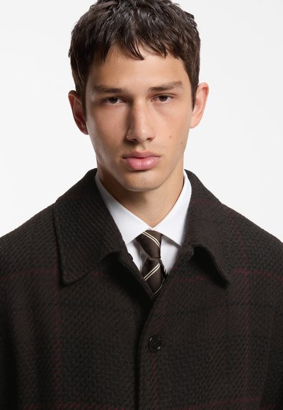 Jeune homme aux cheveux courts et foncés portant un manteau sombre à motifs, une chemise blanche et une cravate rayée sur un fond uni.