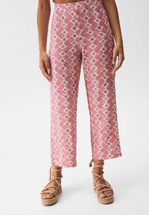 Kobieta ubrana w różowe, szerokie spodnie z wysokim stanem i wzorem oraz beżowe sandały na platformie owinięte liną, stojąca na białym tle.