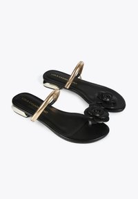 Sandalias planas negras con un acento floral, herrajes metálicos dorados y una suela moldeada. Presenta una tira ancha y un diseño de punta abierta.