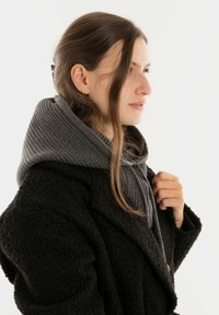 Jonge vrouw met lang bruin haar in een zwarte textured jas en een grijze gebreide sjaal met capuchon, die naar rechts kijkt tegen een eenvoudige lichte achtergrond.