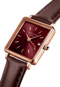 Montre bracelet carrée avec un cadran bordeaux, des aiguilles dorées et un guichet de date, montée sur un bracelet en cuir marron texturé.