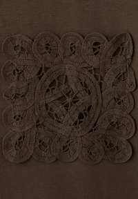 Dunkelbrauner Stoff mit einem kunstvoll bestickten Spitzenquadrat in der Mitte, das Schlaufen und kreisförmige Muster zeigt.