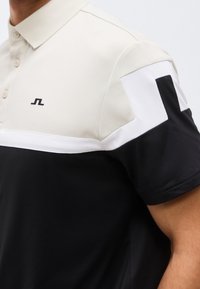 Κοντομάνικο πόλο πουκάμισο με μπλοκ χρωμάτων σε μαύρο, άσπρο και μπεζ, και ένα μικρό μαύρο λογότυπο στο στήθος.