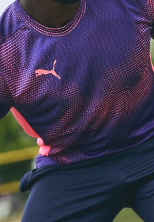 Persoon draagt een paars-roze Puma sportshirt met een patroon en donkere broek, in een actieve houding buiten.