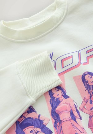 Detailní pohled na bílou mikinu s vroubkovaným límcem a manžetami, zdobenou barevným motivem tří animovaných žen ve stylizovaných outfitech.