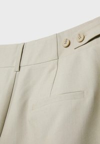 Gros plan sur la ceinture de pantalon beige avec deux boutons beiges et une couture de poche latérale.