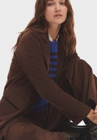 Blazer de pana marrón sobre un suéter a rayas azules, combinado con pantalones marrones. Presenta solapa, bolsillo y detalles de tejido texturizado.