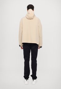 Beige hooded jacket with a loose fit, cropped hem, and black striped accents at cuffs, paired with dark pants and white sneakers.