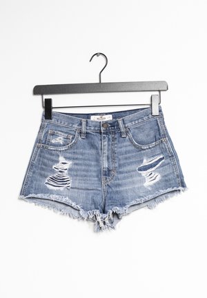 Shorts de mezclilla azul desgastada con dobladillos desflecados colgados en una percha negra contra un fondo blanco.