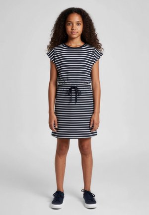 Ragazza giovane con lunghi capelli ricci, indossa un vestito a righe blu navy e bianco con cintura in vita e sneakers blu navy, in piedi su uno sfondo semplice.