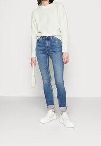 Suéter esponjoso de color verde claro, jeans de mezclilla azul ajustados, con los tobillos remangados, y zapatillas blancas, sosteniendo un pequeño bolso blanco.