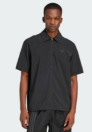 Svart zip-up polo skjorta med spetsig krage, korta ärmar och en subtil logotyp på vänster bröst. Materialet verkar lätt och slätt.