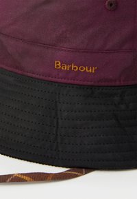 Chapeau seau bordeaux avec un bord noir, featuring des coutures jaunes et le mot "Barbour" brodé à l'avant. Tissu texturé.