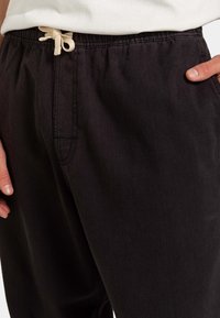 Black denim pants with a tapered design, featuring a drawstring waistband in beige and side pockets. Textured fabric with a smooth finish.